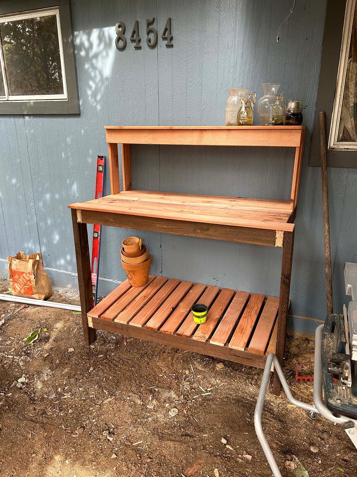 Wooden shelf unit with planter, jug, and small items against a gray exterior wall by the driveway