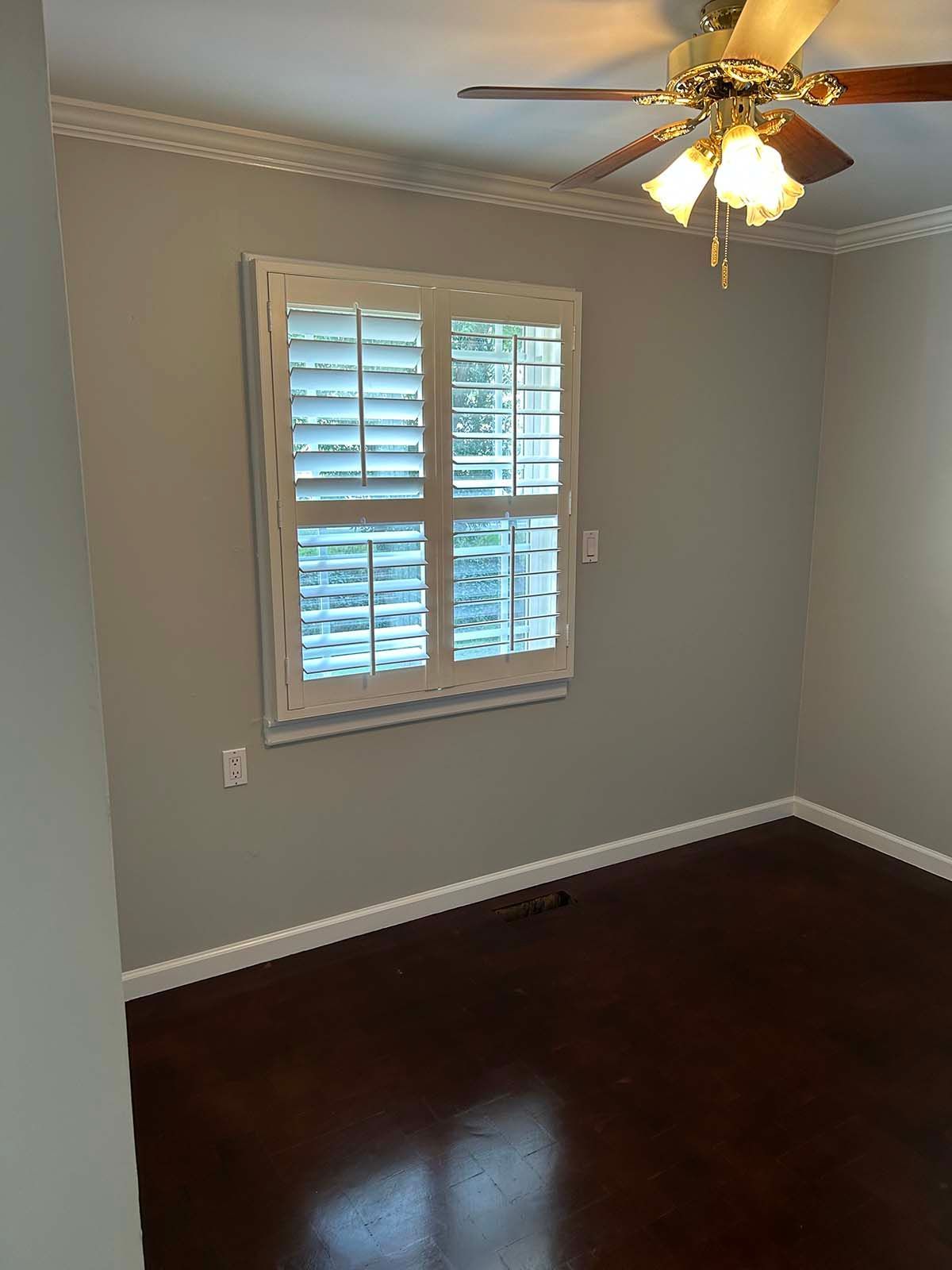 Empty room with gray walls, dark hardwood floor, white window shutters, and a ceiling fan with lights.