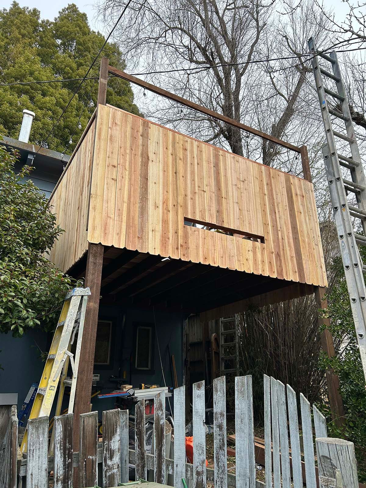 Wood-clad tiny house on stilts with a ladder, surrounded by trees and a metal fence
