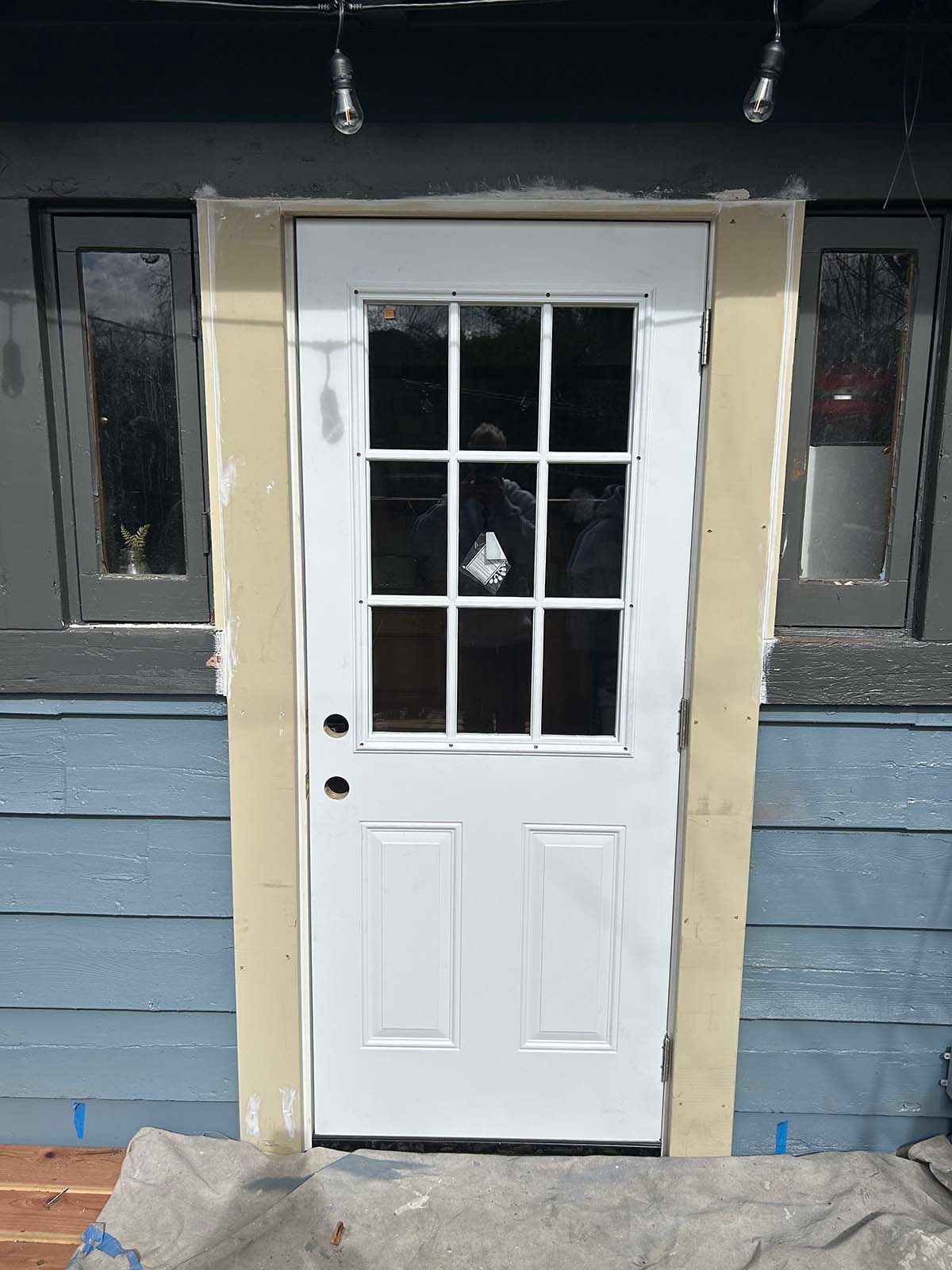 White front door with window panes in blue siding, flanked by two narrow windows on a porch.