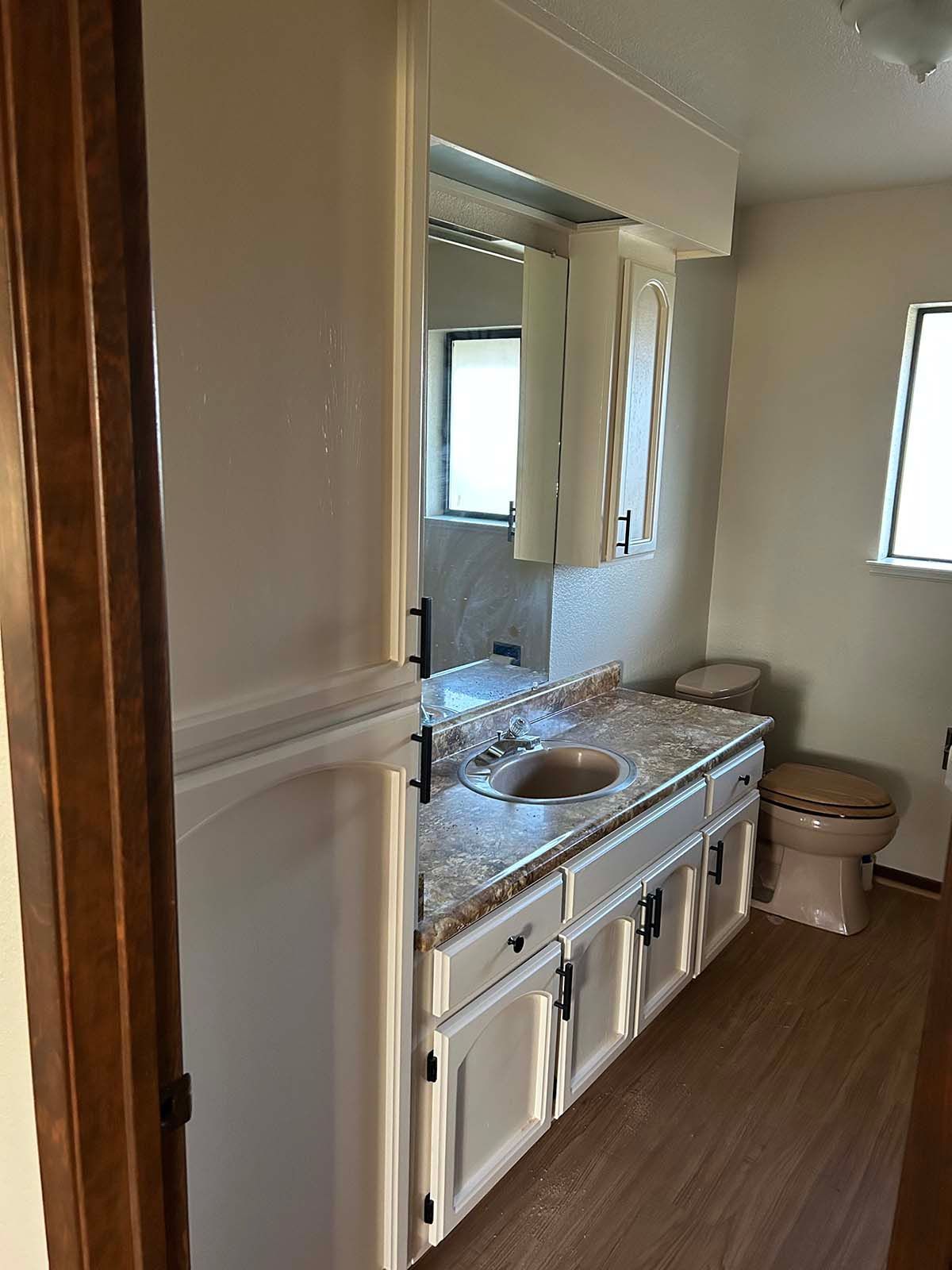 Bathroom vanity with granite countertop, white cabinets, mirror, and a toilet near a window.