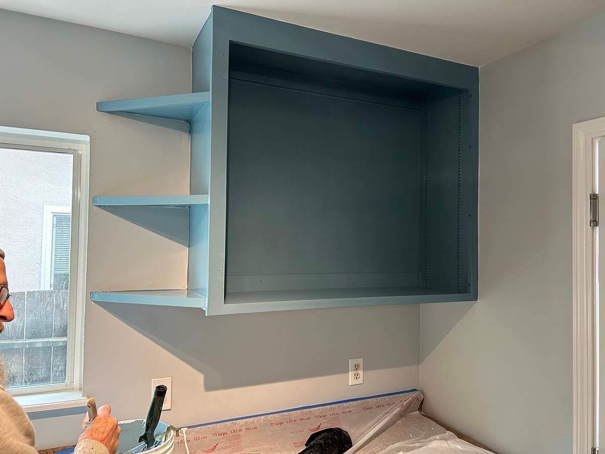 Large blue wall-mounted cabinet with open shelves above a desk in a small room.