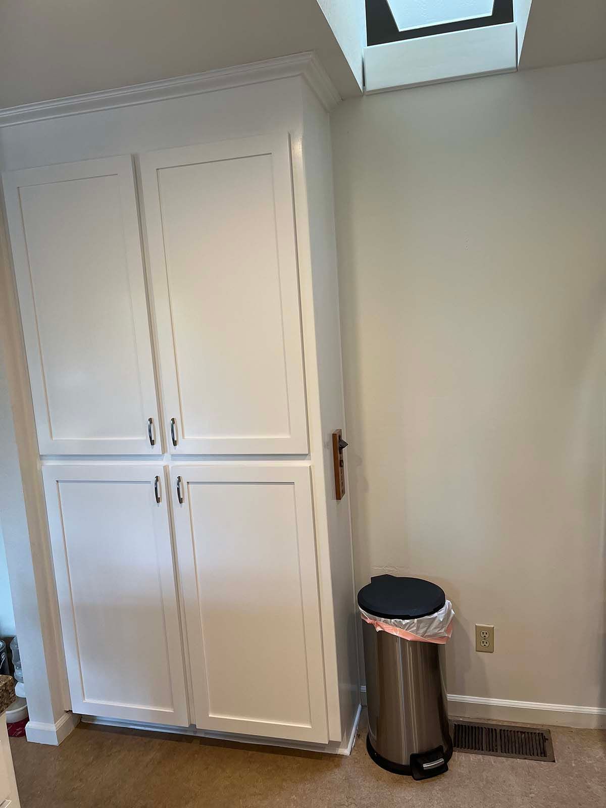 White built-in cabinet beside a stainless trash can in a bright room with a skylight.