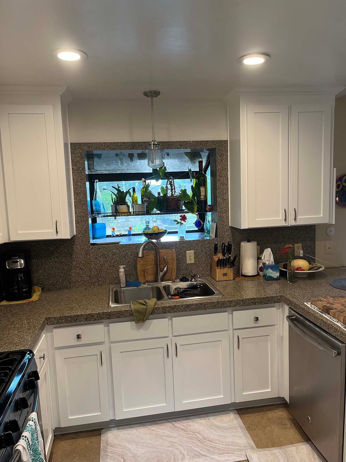 Bright kitchen with white cabinets, granite counters, and a sink beneath a window with hanging plants.
