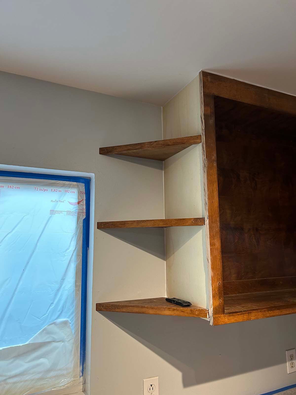 Wooden corner shelves mounted on a beige wall beside a blue-taped window opening.