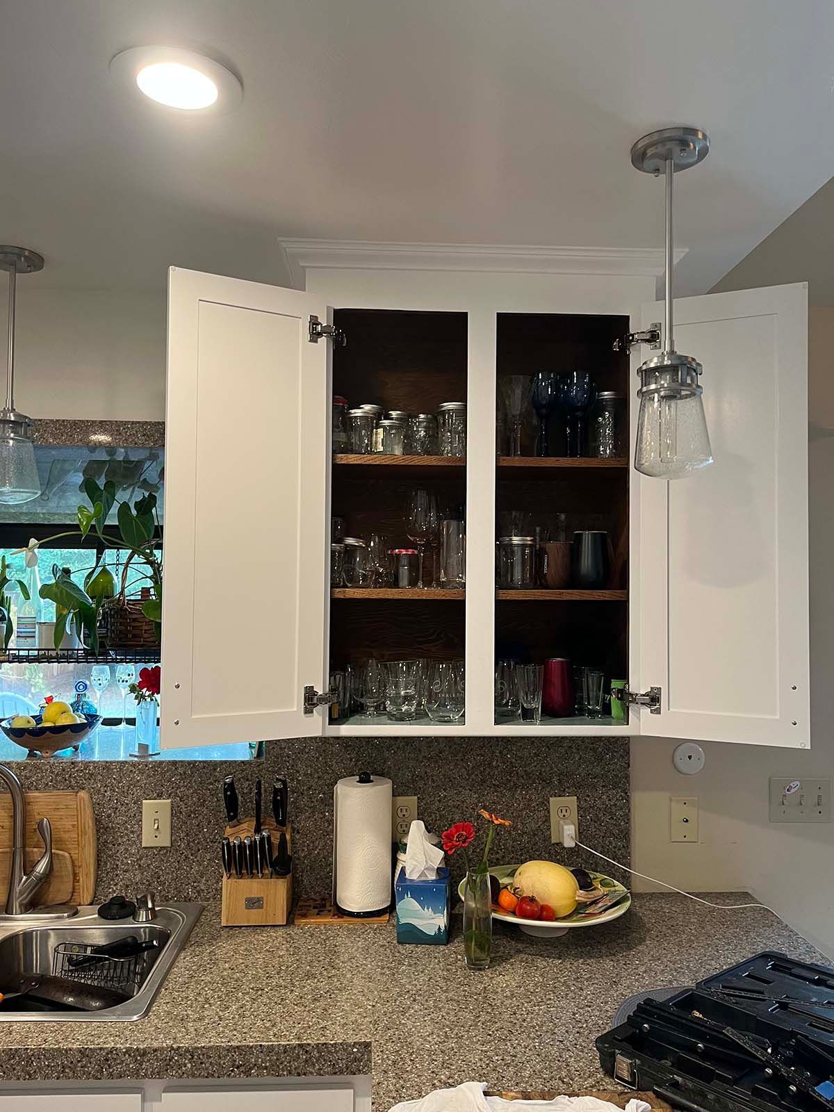 Kitchen with open cabinets, granite countertop, sink, dish soap, fruit bowl, and glassware behind cabinet doors.