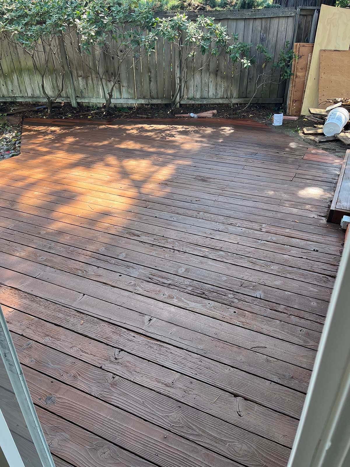 Wooden backyard deck in sunlight, with a weathered fence and garden items along the edge.