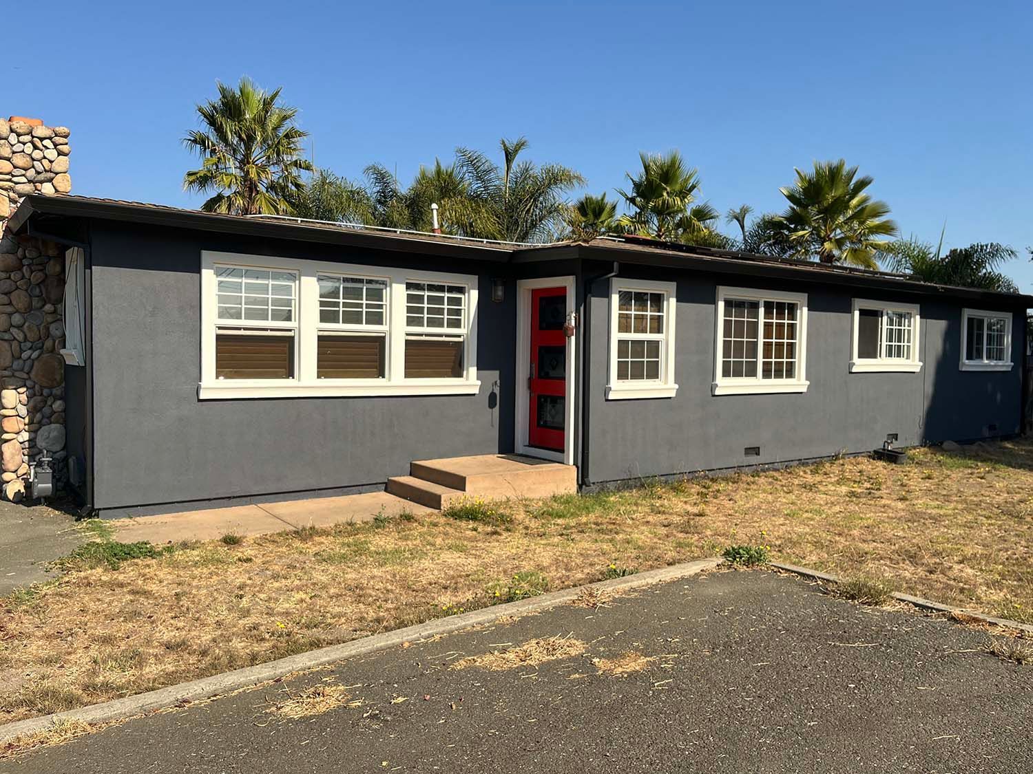 Single-story gray house with white-trimmed windows, red door, and dry front yard under a clear sky