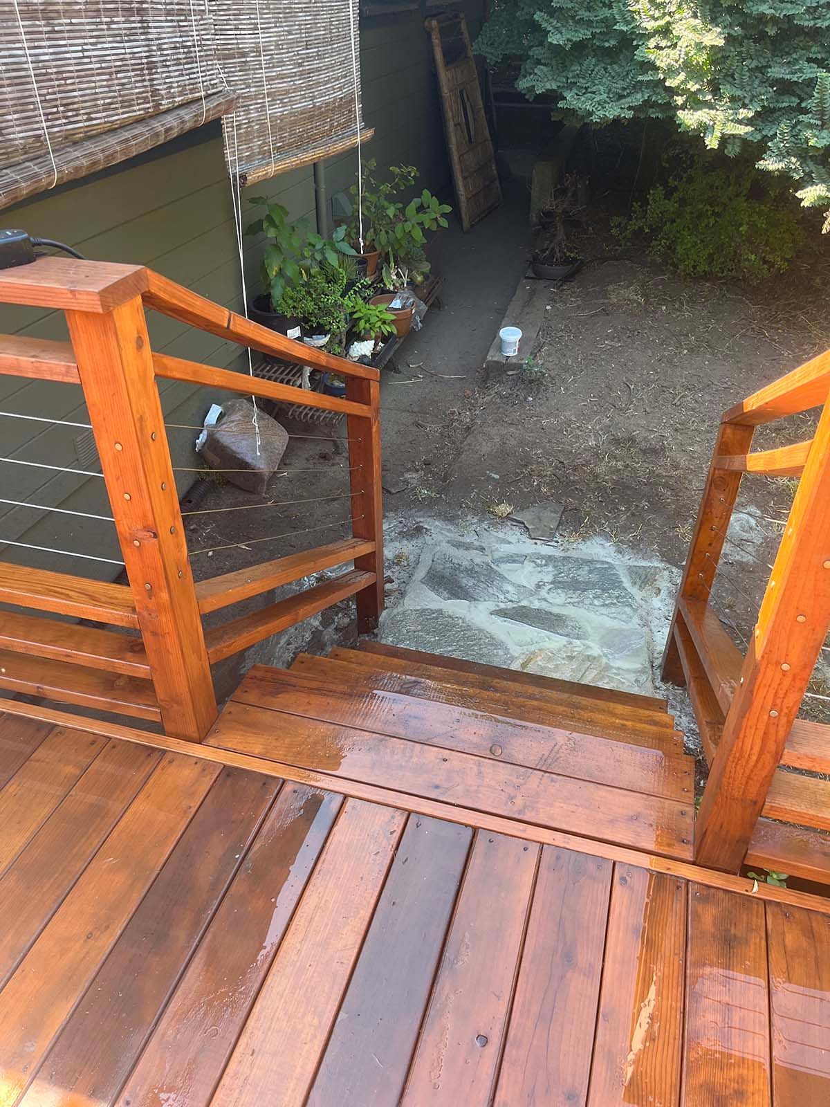 Wooden deck stairs leading down to a small gravel courtyard with plants and a brick wall.