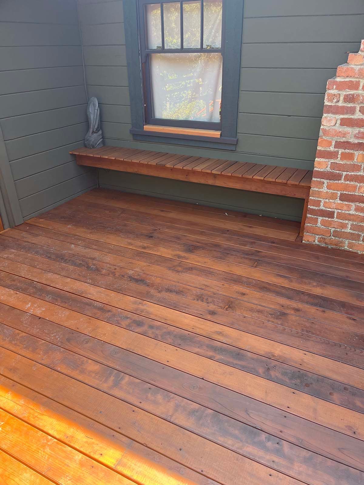 Wooden deck with built-in bench beneath a window, dark siding, and a brick wall edge.