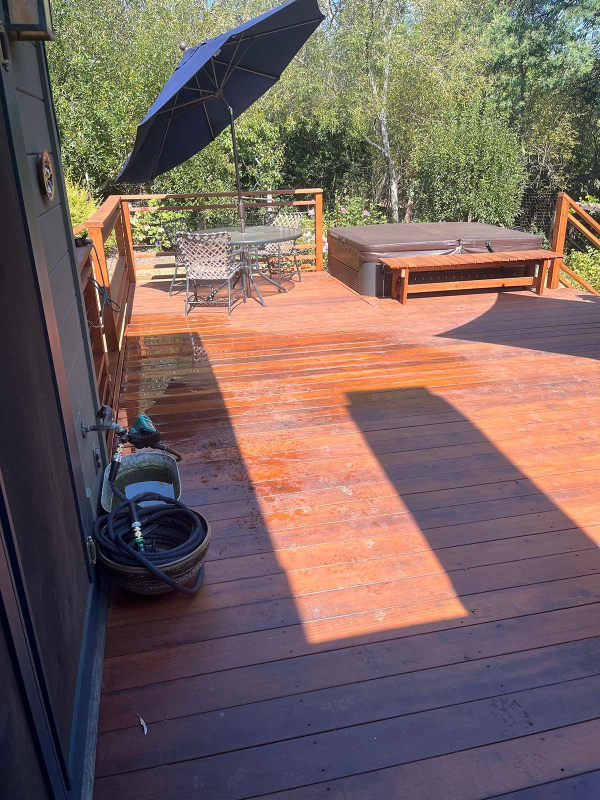 Wooden deck with umbrella, patio table, lounge chair, and coiled hose in bright sunlight.