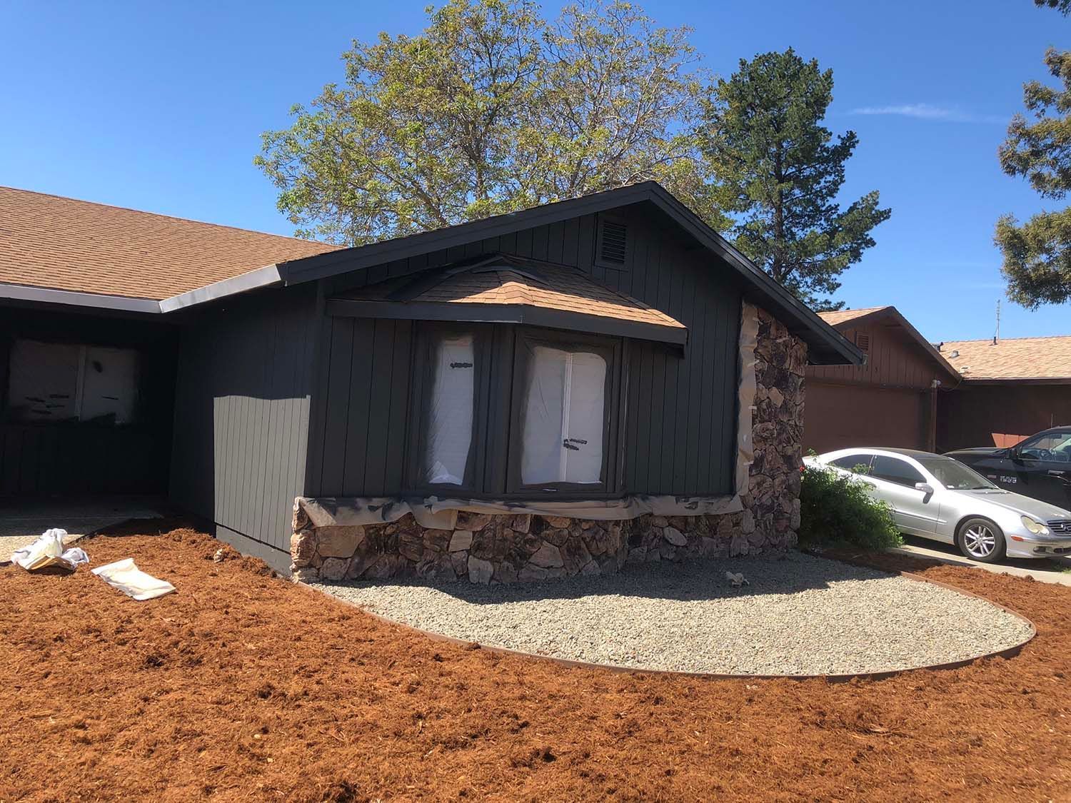 Single-story house exterior with dark siding, stone base, and a white car in the driveway under a blue sky