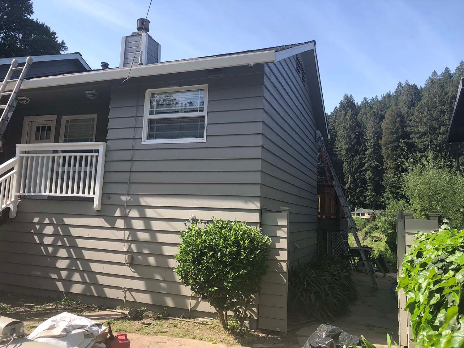 Gray house exterior with white trim, small porch, and a narrow path beside tall trees and bushes