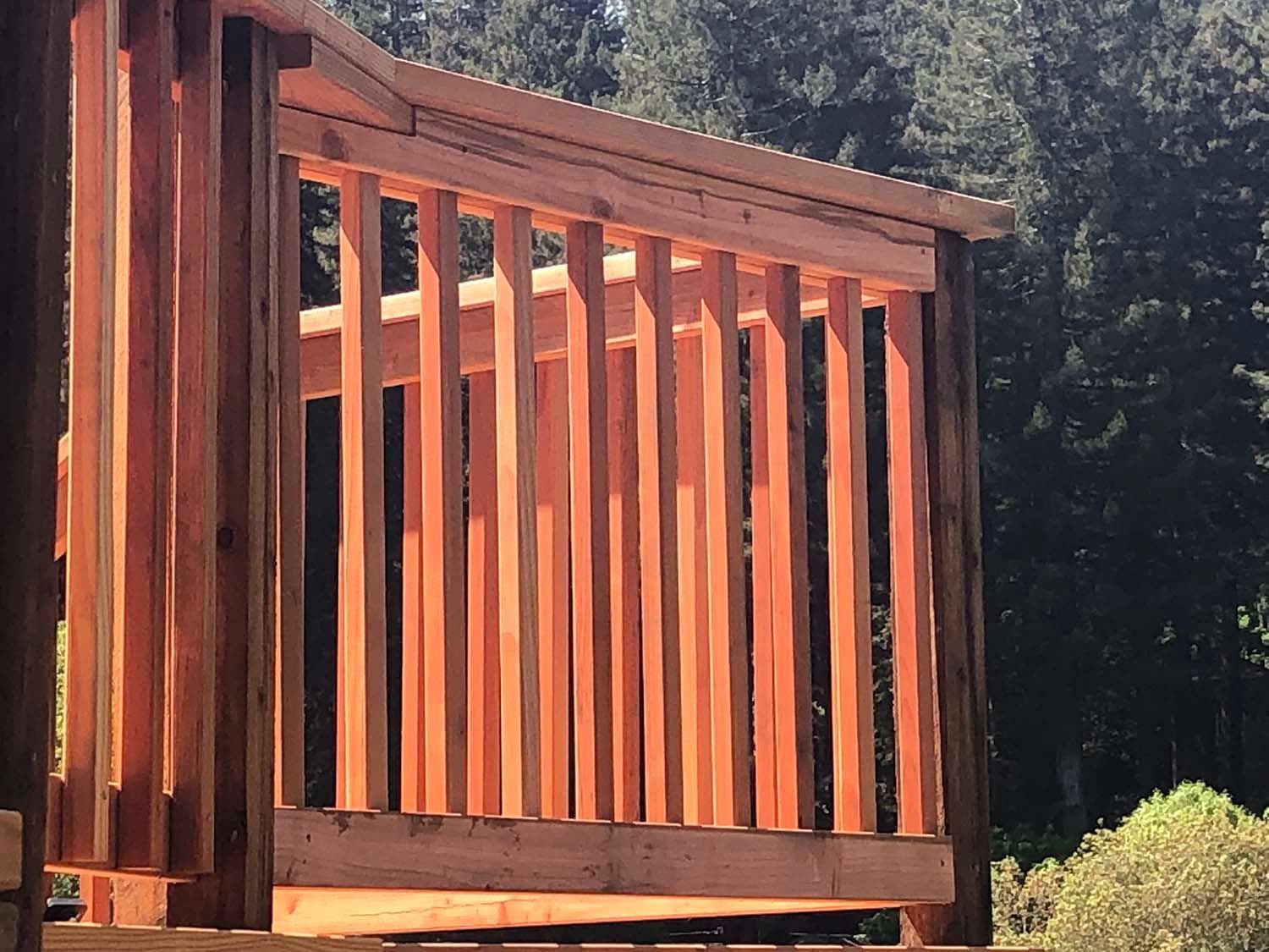 Wooden deck railing with vertical slats beside a forested hillside in warm sunlight