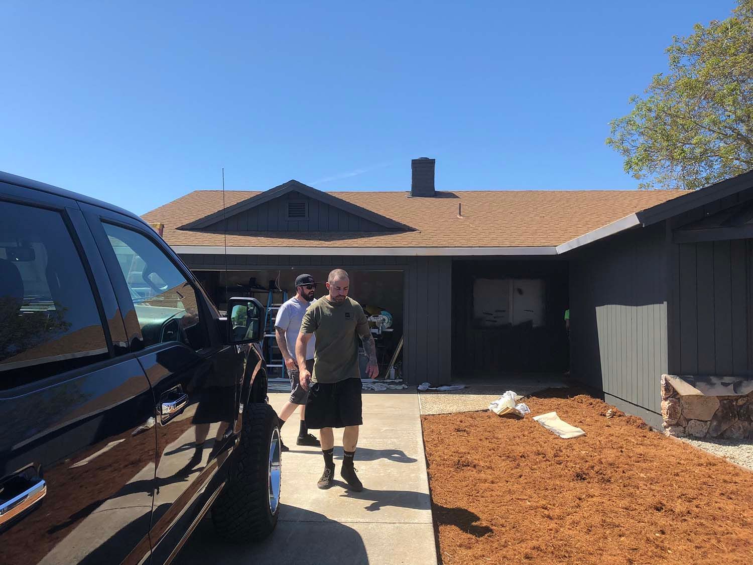 Person standing in a driveway beside a black truck in front of a house under a clear blue sky