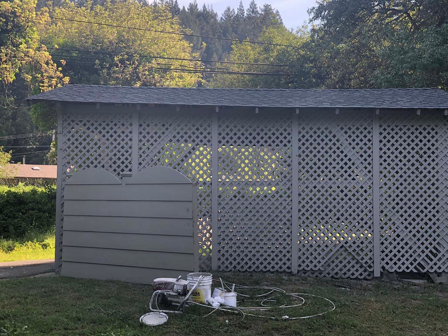 White lattice garden shed with a sloped roof and lawn equipment in front, set against trees.