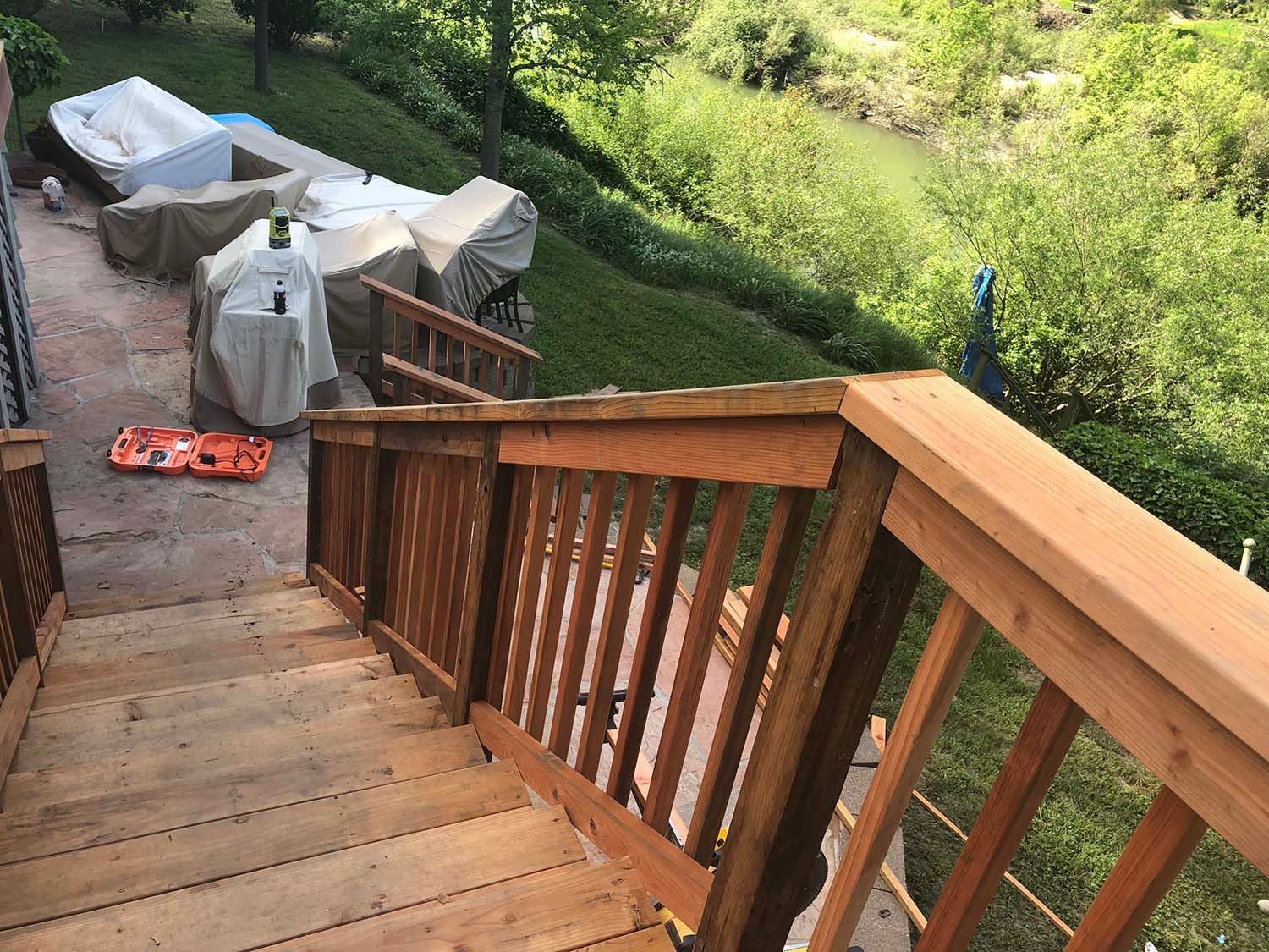 Wooden deck stairs descending to a patio with covered furniture and a green yard beyond