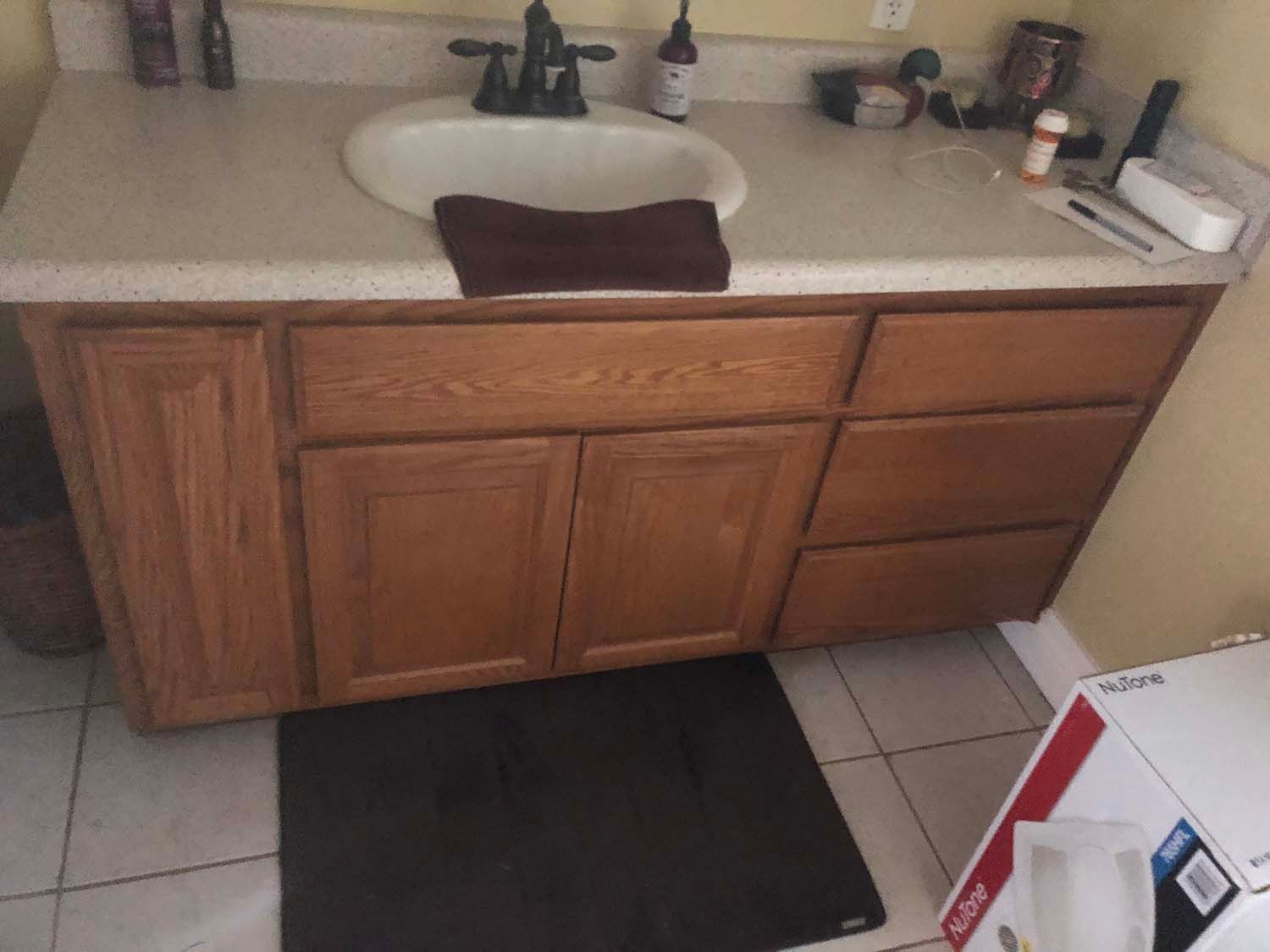 Bathroom vanity with sink, wooden cabinets, and a black bath mat on white tile floor.