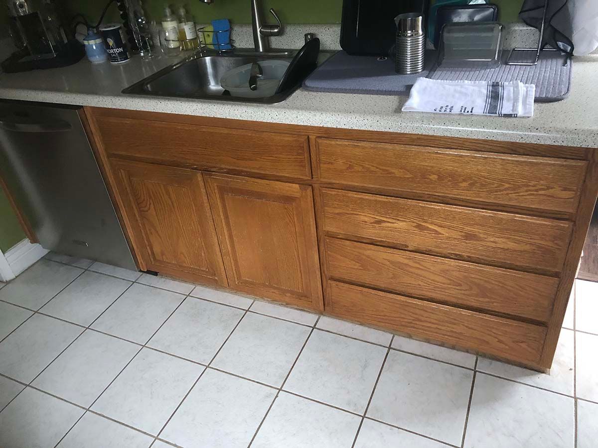 Wooden kitchen cabinets and sink beneath a speckled countertop with white tiled floor.