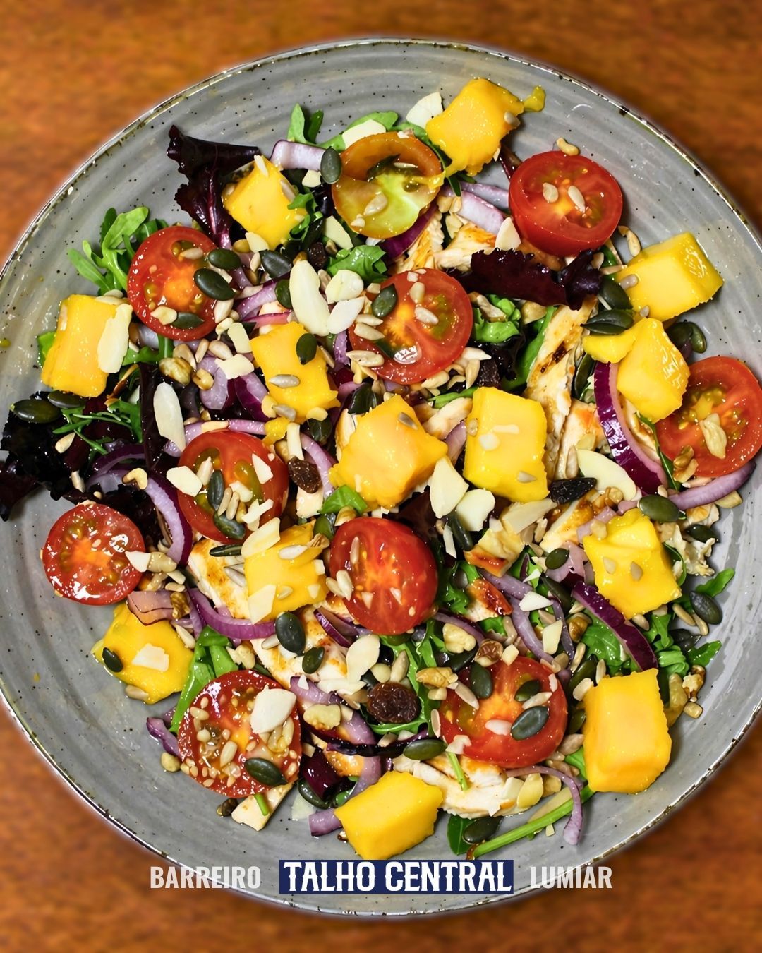 Salada colorida com tomates-cereja, manga, cebola roxa, abacate e sementes em uma tigela cinza.