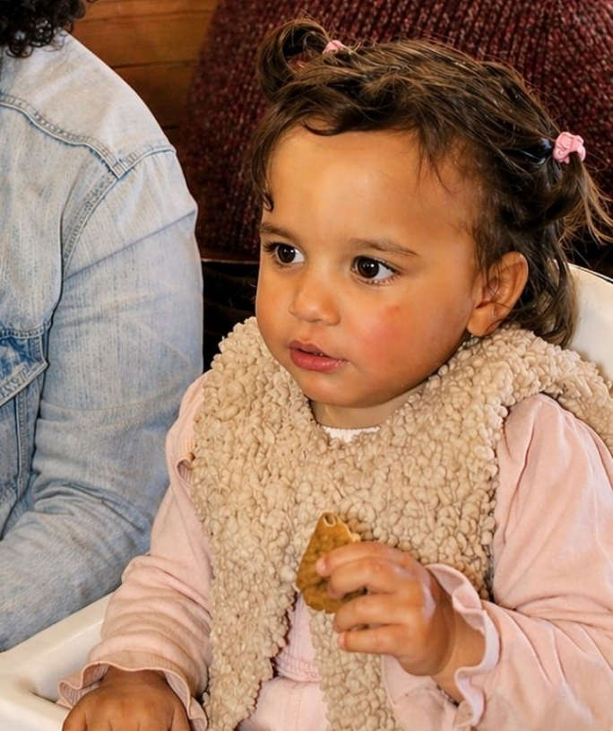 Criança pequena com camisa rosa e colete bege segurando um lanche, sentada dentro de casa ao lado de um adulto de calça jeans.