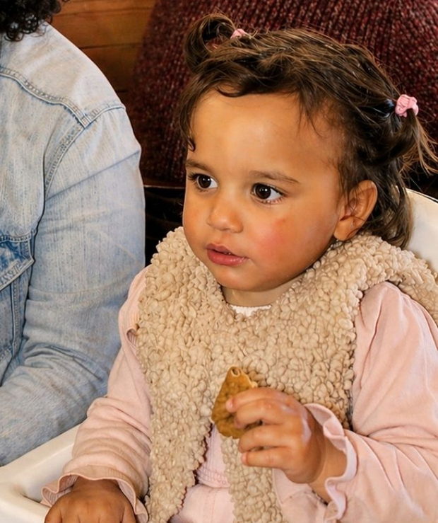 Criança pequena com camisa rosa e colete bege segurando um lanche, sentada dentro de casa ao lado de um adulto de calça jeans.