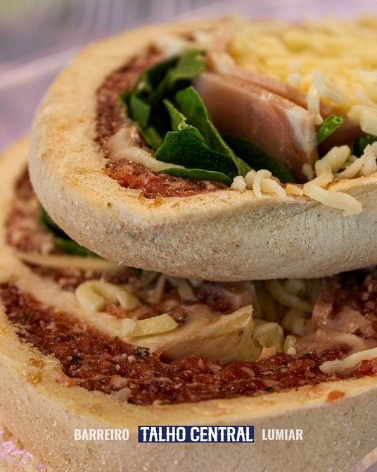 Imagem em close-up de um pão achatado recheado com queijo ralado, verduras e carne, servido em um prato com a marca da empresa.