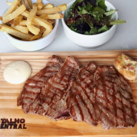 Bife grelhado com batatas fritas, salada e molho servido em uma tábua de madeira com pão torrado.