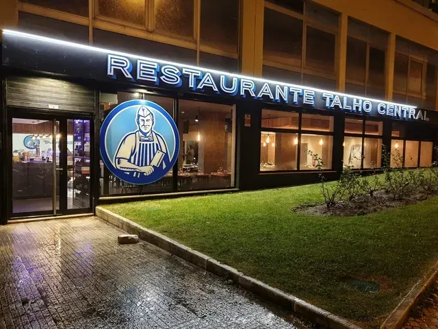 Vista externa noturna do Restaurante Tairo Central, com letreiro iluminado, passarela molhada e entrada gramada.