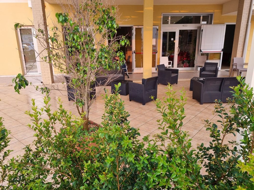Patio con sedie in vimini nere, porta a vetri e arbusti verdi di fronte a un edificio beige.