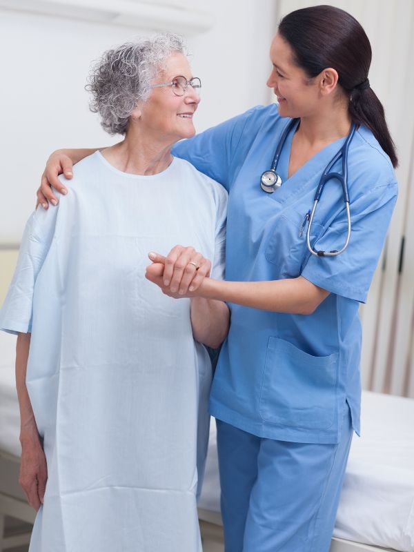 Un'infermiera in camice blu sorride e tiene per mano un paziente in camice bianco in una stanza della clinica.