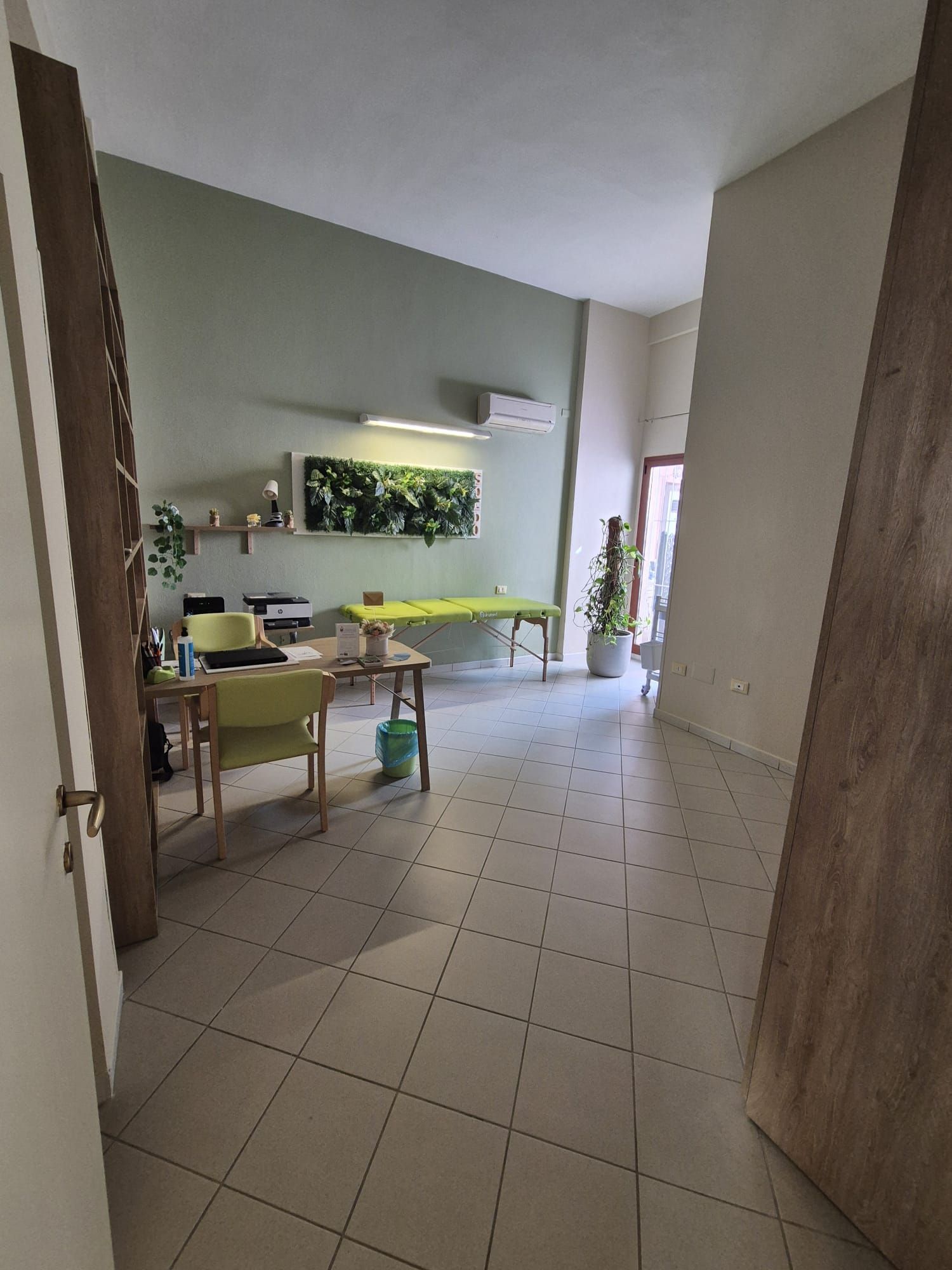 Tranquilla stanza interna con pavimento piastrellato, lungo tavolo, mensole a muro e piante vicino a una porta luminosa.