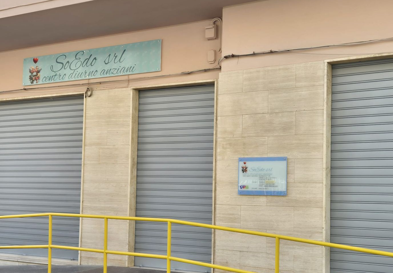 Negozio chiuso con serrande metalliche, ringhiera gialla e insegna turchese sopra l'ingresso