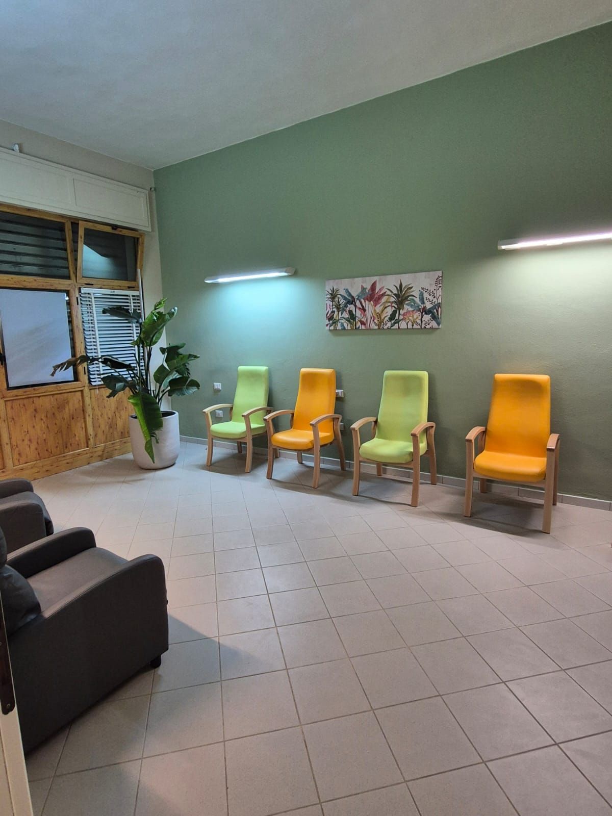 Sala d'attesa con pareti verdi, pavimento piastrellato e sedie colorate accanto a una finestra con una pianta in vaso.