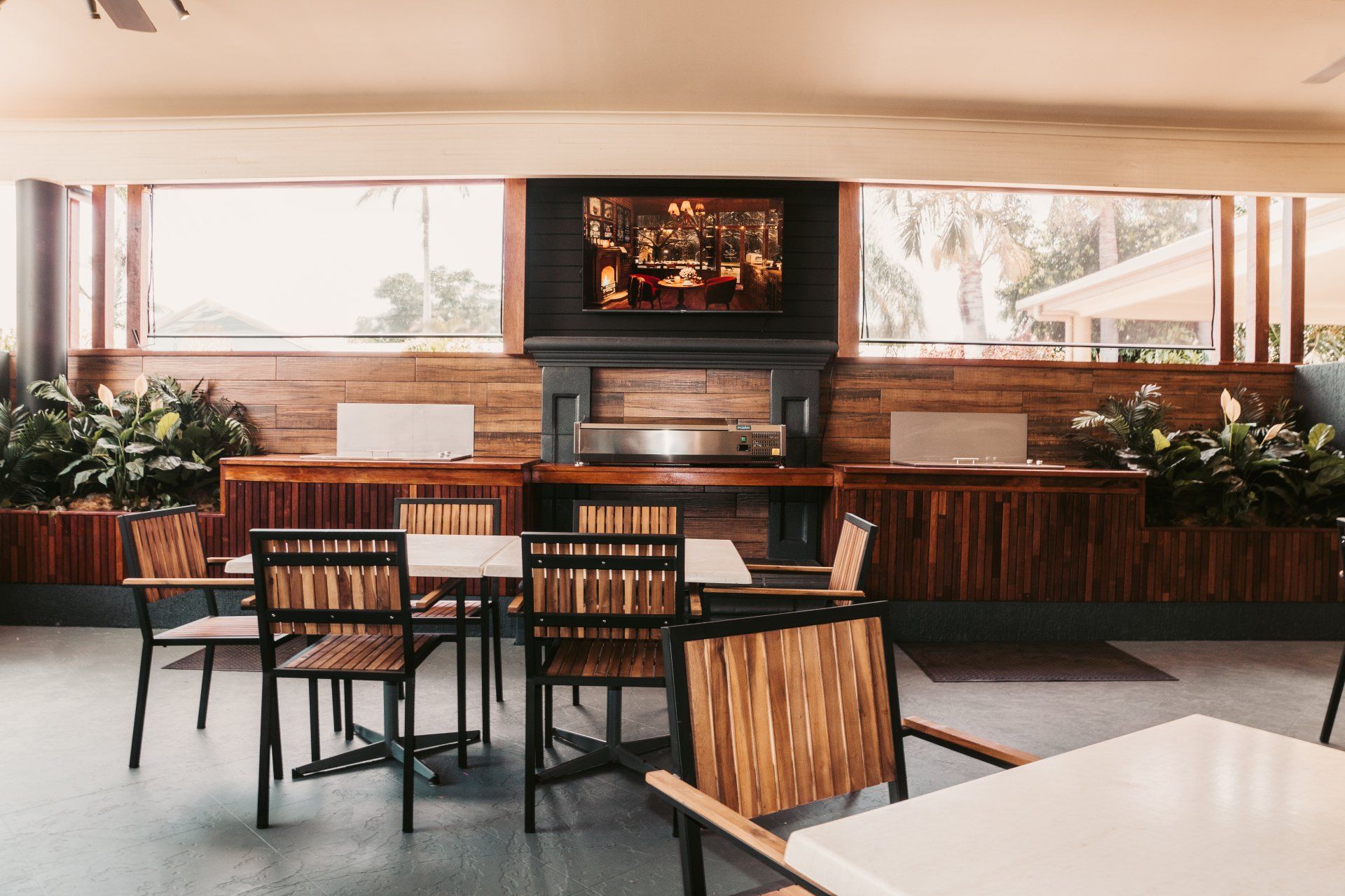 Modern lounge with wooden tables and chairs, bar counter, potted plants, and a wall-mounted TV by large windows
