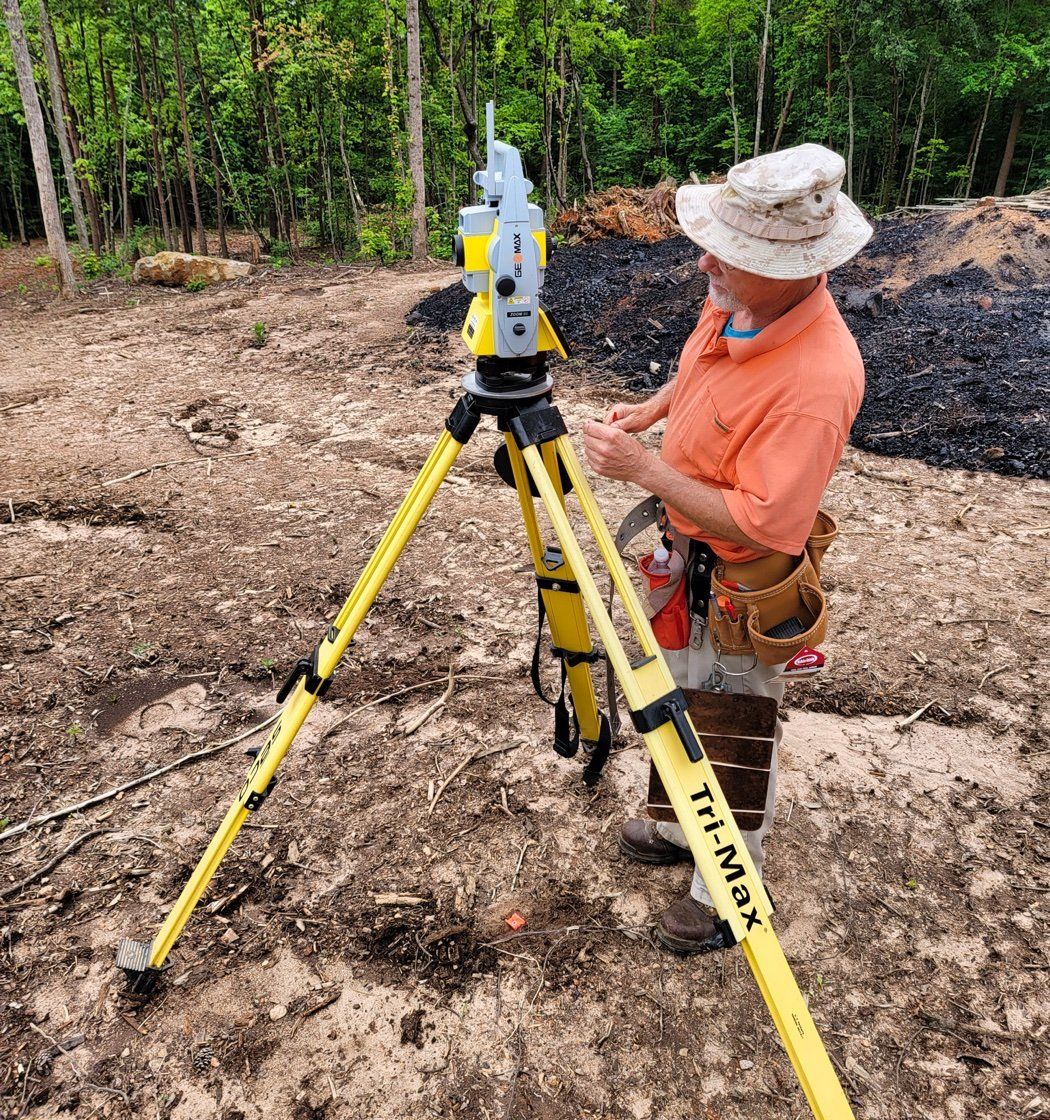 A surveyor in an orange shirt is standing next to a yellow tripod.
