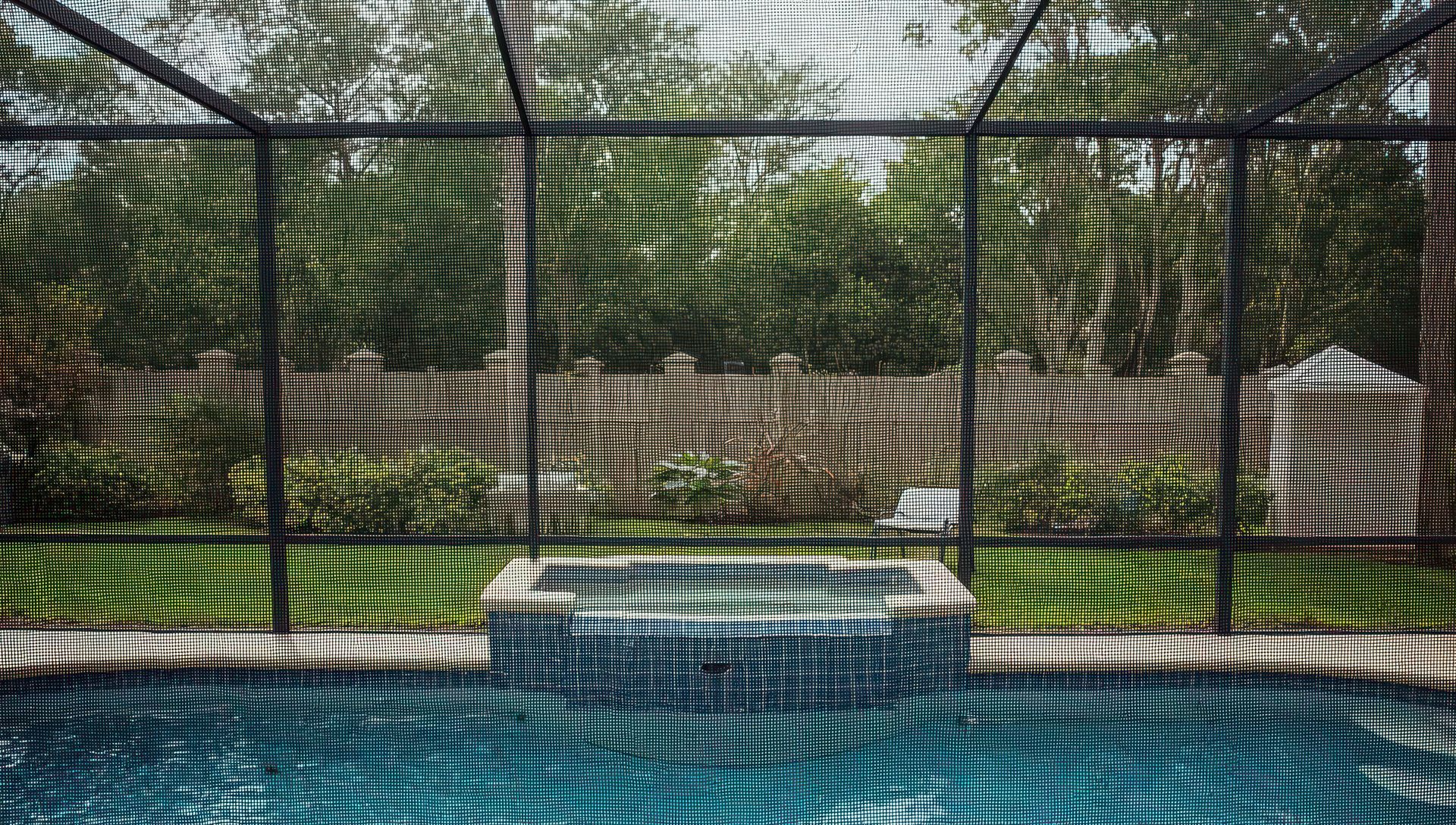 A swimming pool and tiled hot tub viewed through a screened lanai with dappled light casting shadows on the pool water.