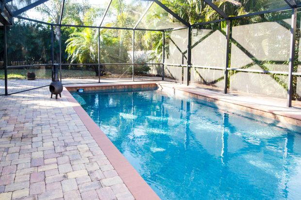 A swimming pool with blue water enclosed by a screened lanai, surrounded by light-colored pavers.