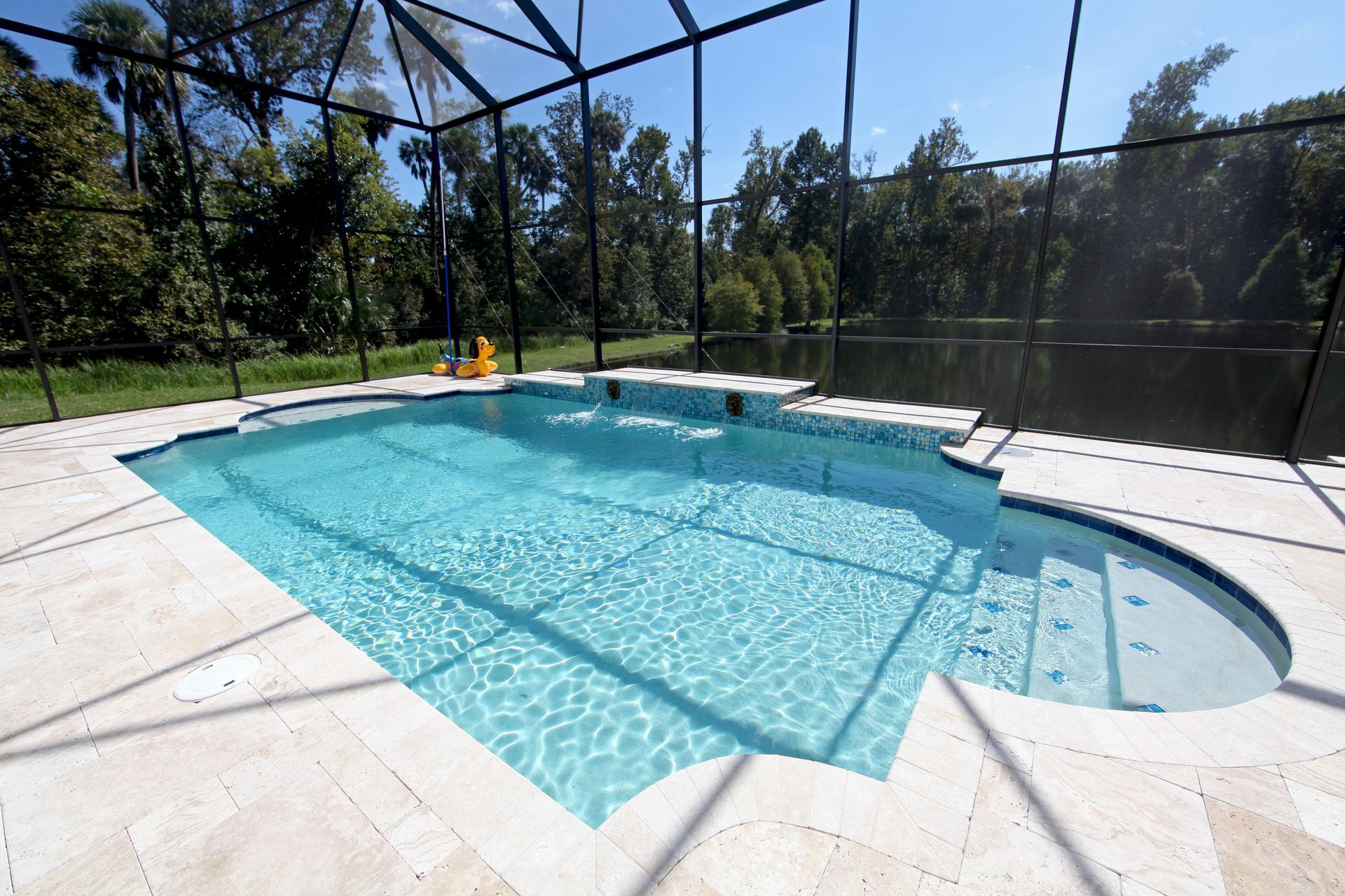 A screened-in backyard swimming pool with light blue water, a built-in step area, and tan stone decking overlooking trees.