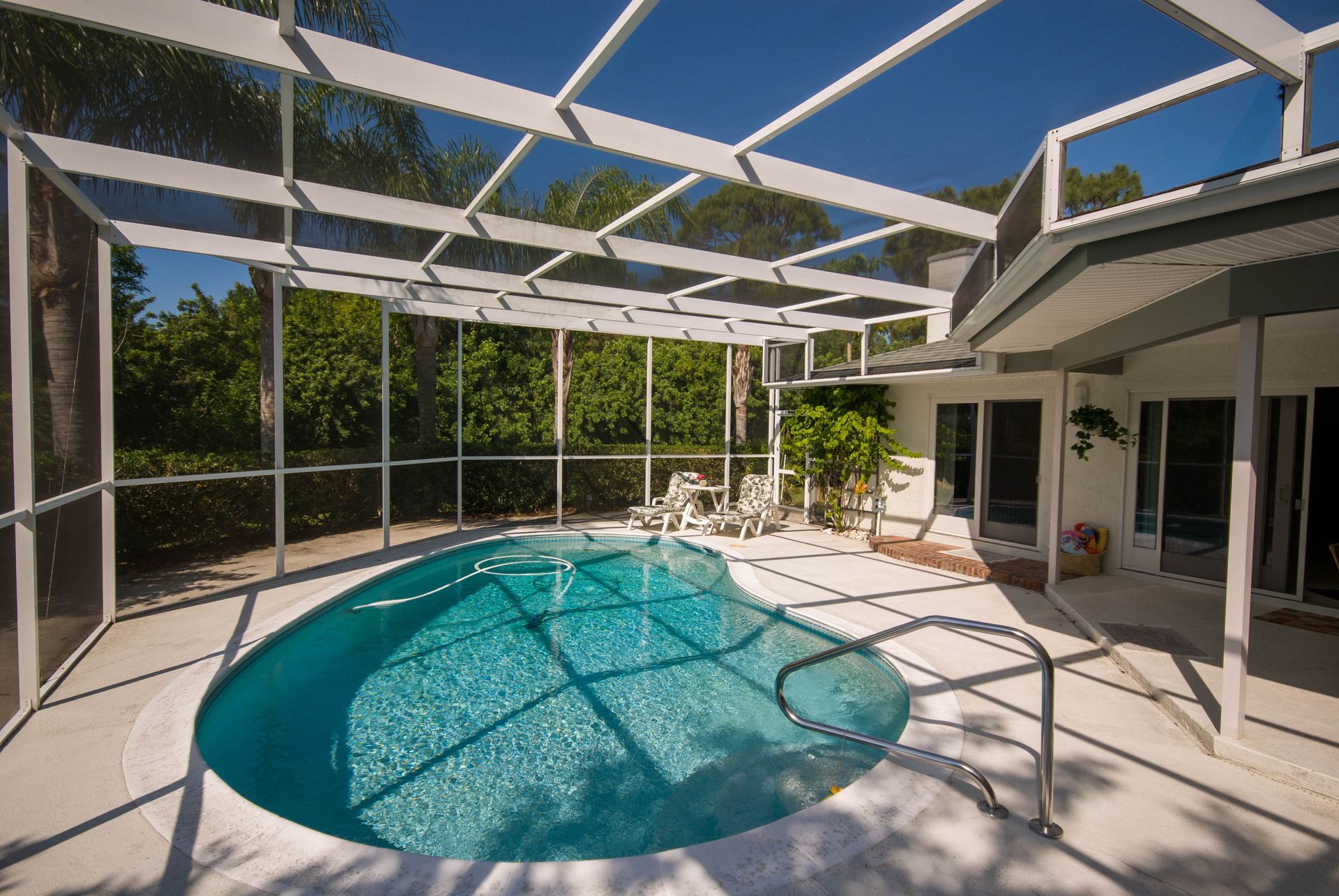 A backyard swimming pool with a turquoise interior, enclosed by a white screen cage, next to a house patio.