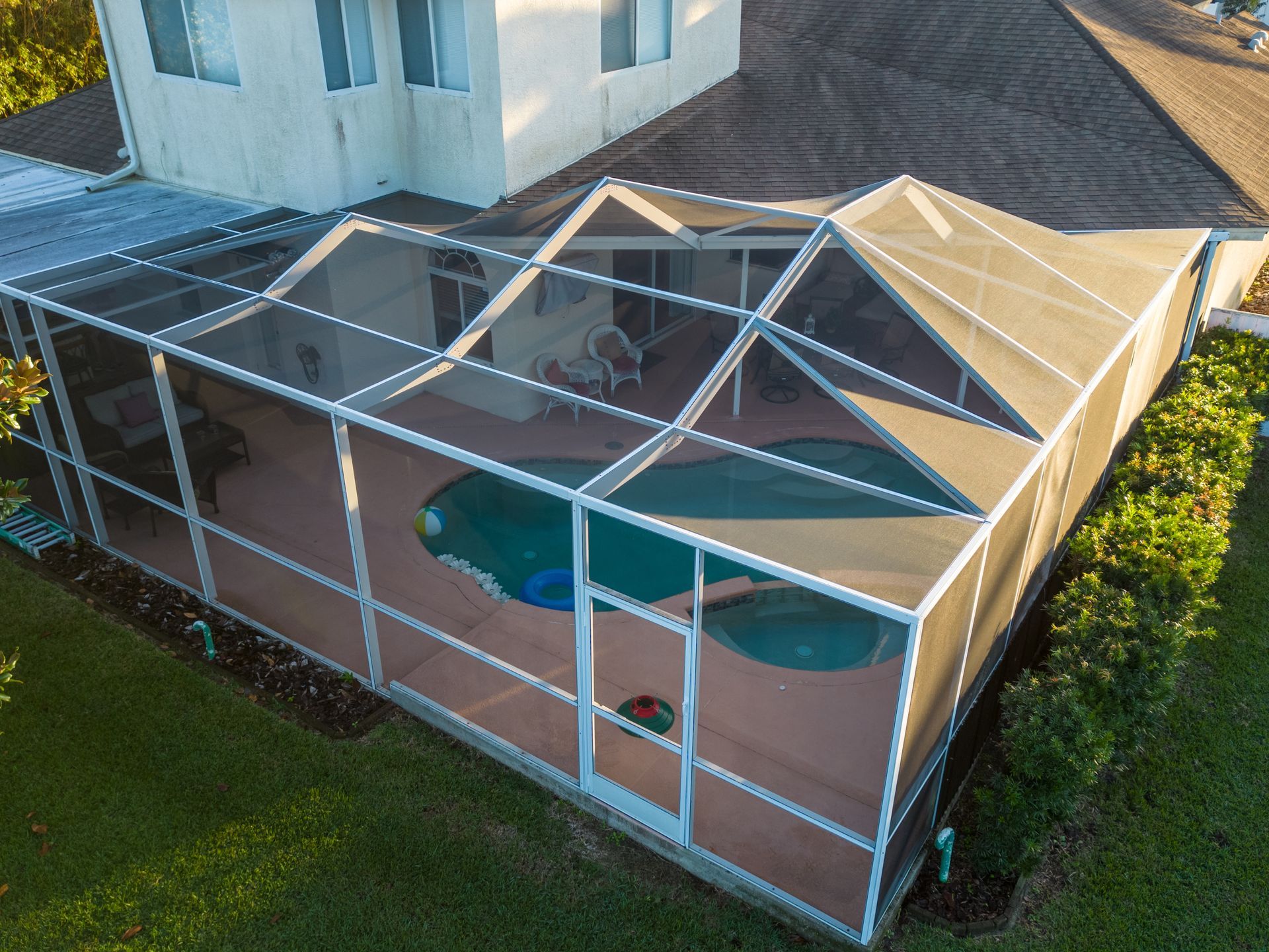 An aerial view of a swimming pool and patio enclosed by a white-framed screen cage attached to a multi-story house.
