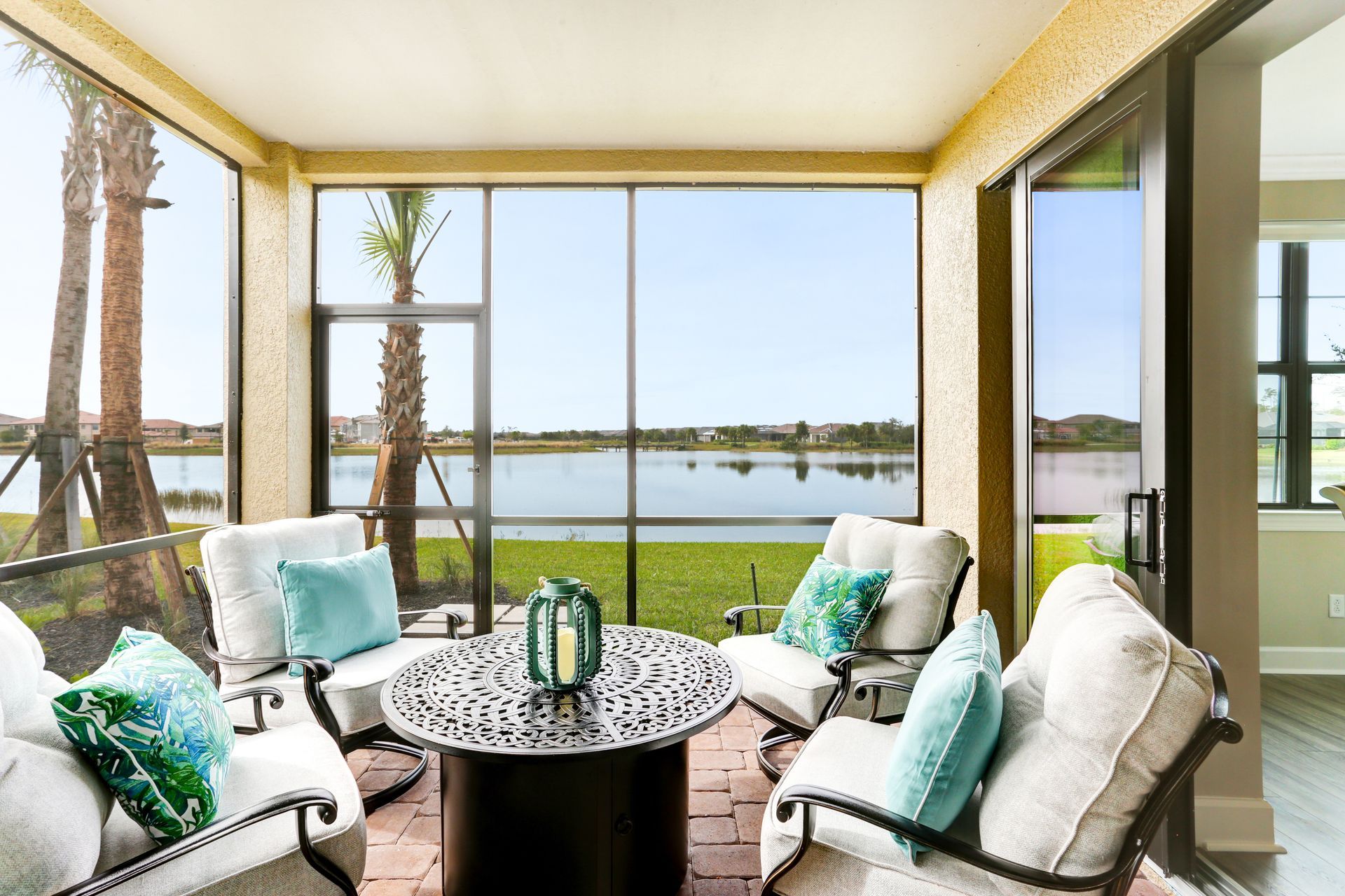 A screened-in patio with four cushioned chairs and a round metal table overlooking a lake and palm trees. A screened-in patio with four cushioned chairs and a round metal table overlooking a lake and palm trees.