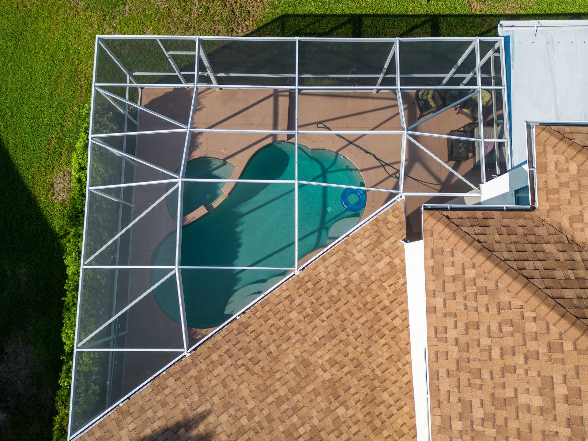 An aerial view shows a backyard swimming pool enclosed by a screened-in white metal cage next to a brown shingled roof.