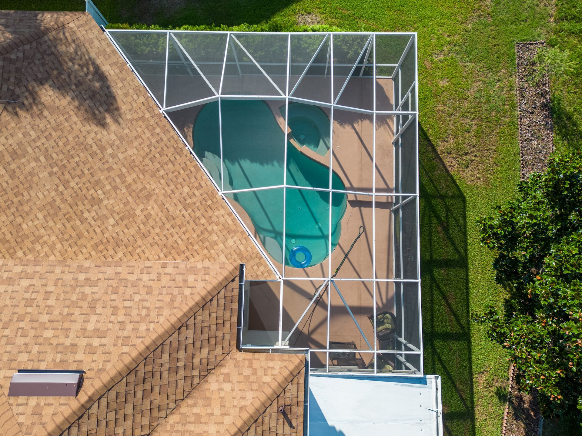 An aerial view of a backyard swimming pool enclosed by a white screen cage, attached to a house with a shingled roof.