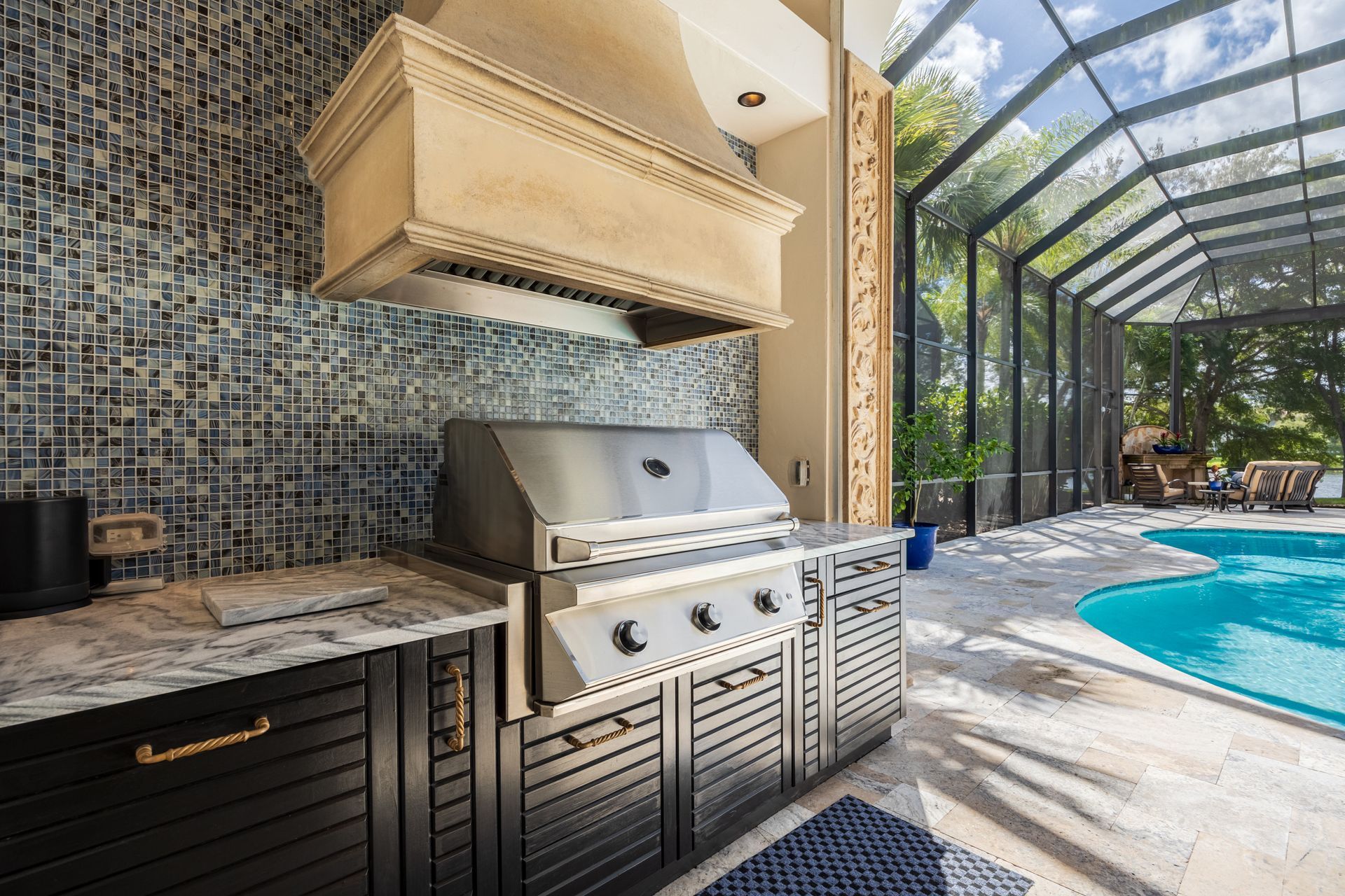 Outdoor kitchen with a stainless steel grill, a tan range hood, black cabinets, and a mosaic tile wall by a pool. Outdoor kitchen with a stainless steel grill, a tan range hood, black cabinets, and a mosaic tile wall by a pool.