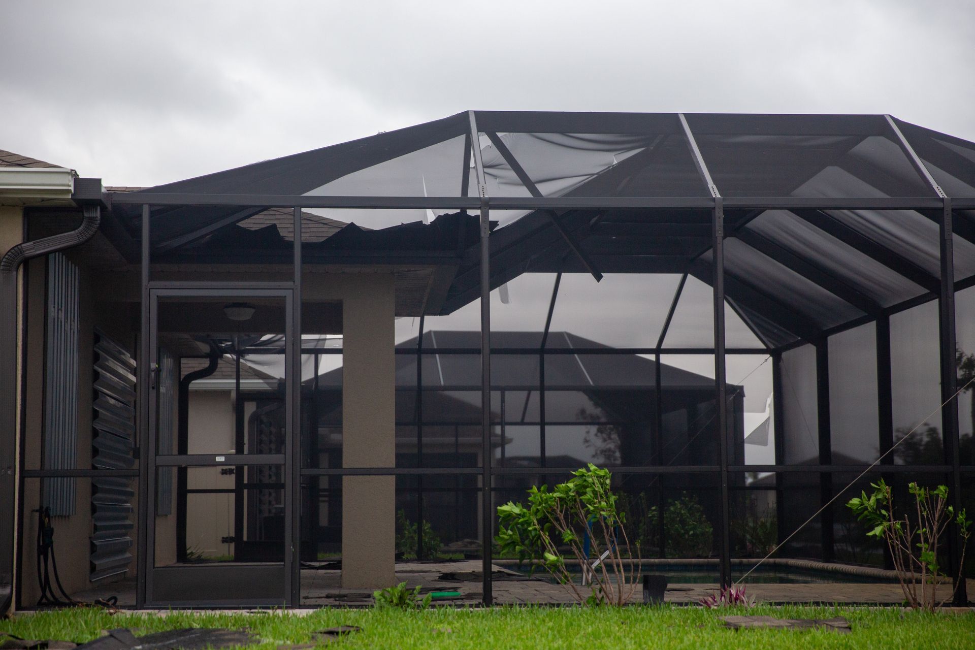 A damaged black aluminum pool screen enclosure attached to a house under a cloudy sky.