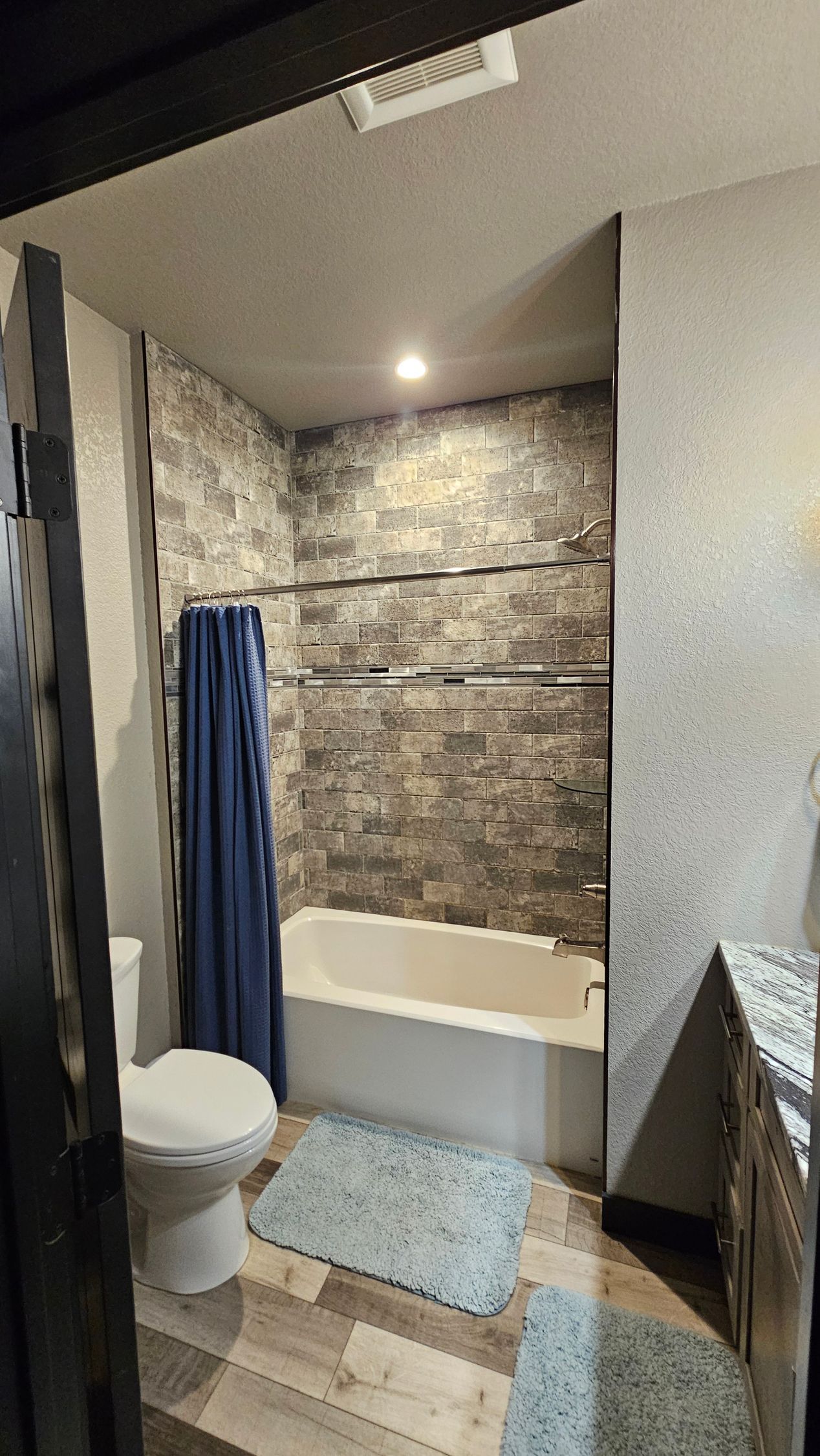 Bathroom with gray stone shower wall, white tub, blue shower curtain, and gray rug.