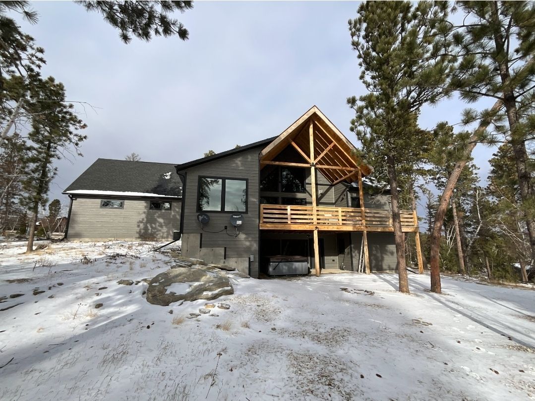 Snowy modern house with a wooden deck, surrounded by pine trees.
