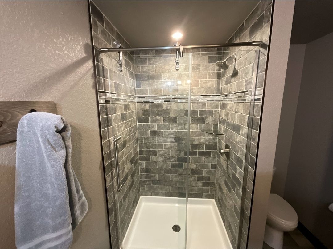 Shower with gray tiled walls, glass doors, and white base. A towel hangs on the left.
