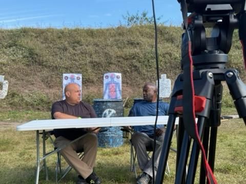 Two men sit at a table outdoors with targets in the background, filming an interview; a tripod is in the foreground.